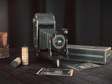 A folding bellows camera on a wooden table with film boxes and loose prints